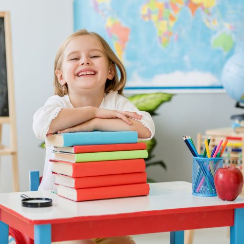 little-smiling-blond-girl-sitting-white-desk-holding-hands-books-spacious-school-class (2)
